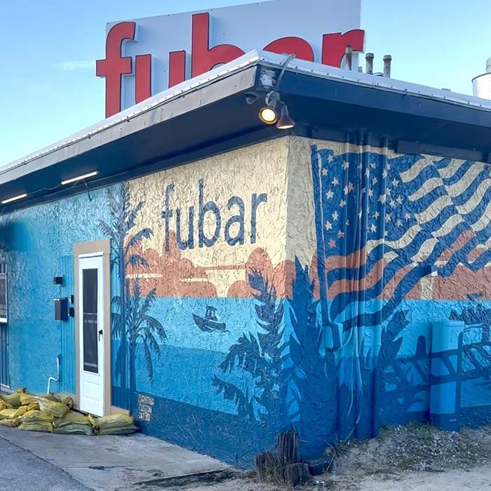 A building with FUBAR Pizza Bar sign, mural of a beach sunset, palm trees, and a waving US flag.